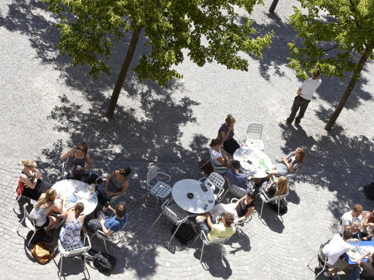 Menschen sitzen draussen an Tischen unter Bäumen.