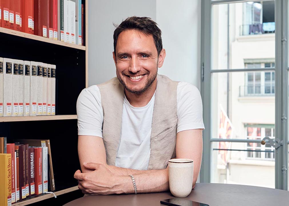 Ein lächelnder Junger Mann neben einem Bücherregal und Kaffeetasse auf dem Tisch.