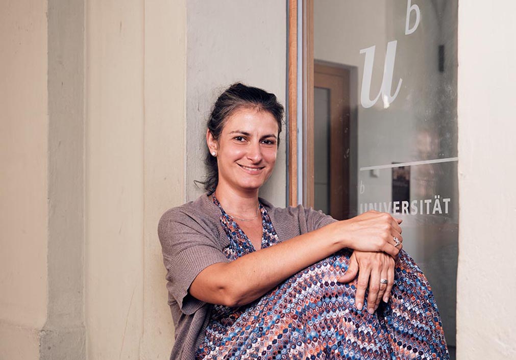 Eine Frau sitzt draussen angelehnt an die Wand, vor einem Fenster mit Uni-Logo.