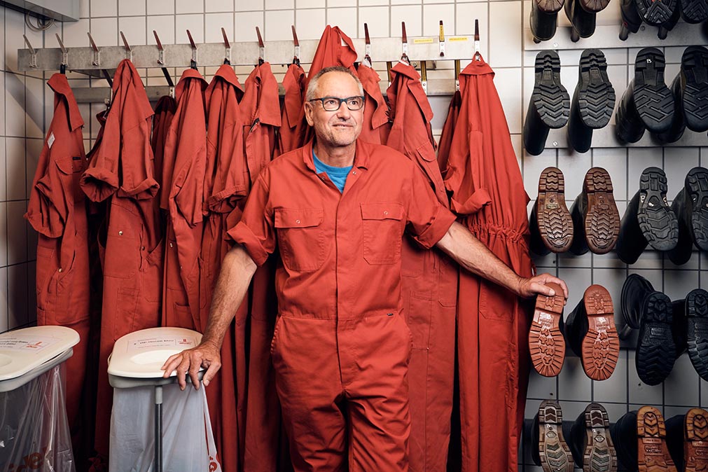 Ein älterer Herr steht in einem Arbeitsoverall und hinter ihm hängen viele Gummistiefel und andere Overalls an der Wand